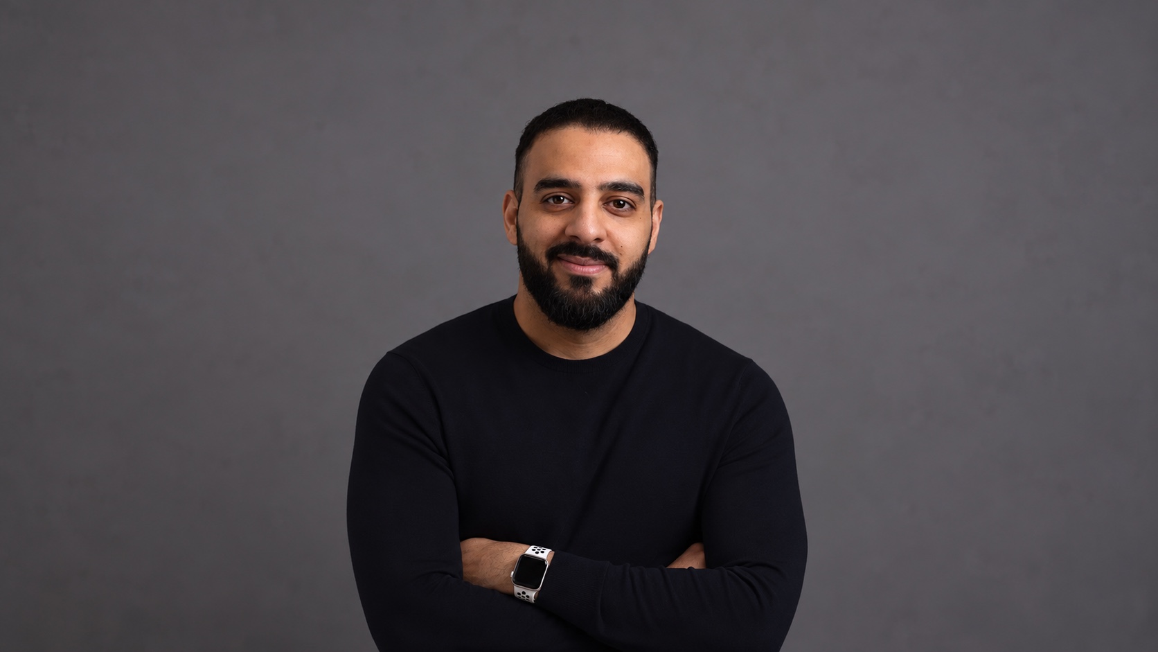 Taher Adel, arms folded, in a black sweater against a grey studio backdrop